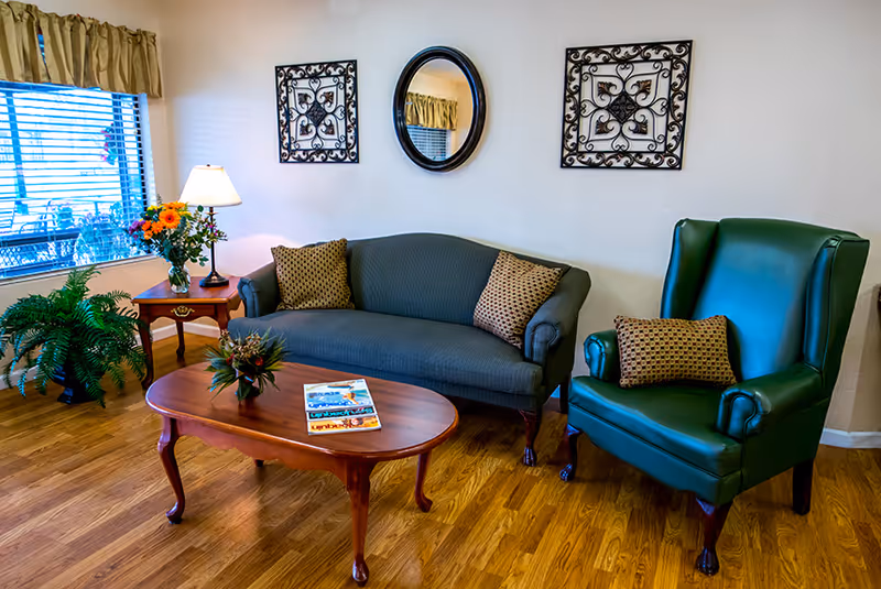 Cozy sitting area with a sofa, green armchair, wooden coffee table, side table with lamp and flowers, wall decor and a window.