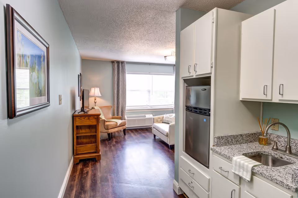 Studio apartment interior with a small kitchenette and sink on the right and a living area with armchairs, TV cabinet, and window at the far end.