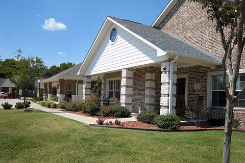 Exterior view of a brick senior living facility building with a covered porch, white columns, and a well-maintained lawn and landscaping under a clear blue sky.
