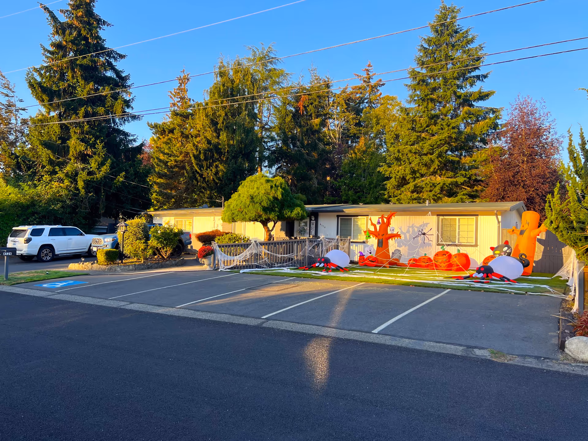Exterior view of a single-story senior care home decorated with Halloween inflatables including pumpkins, spiders, ghosts, and trees with spider webs. There are several tall evergreen trees in the background and a parking area with marked spaces in front of the building.