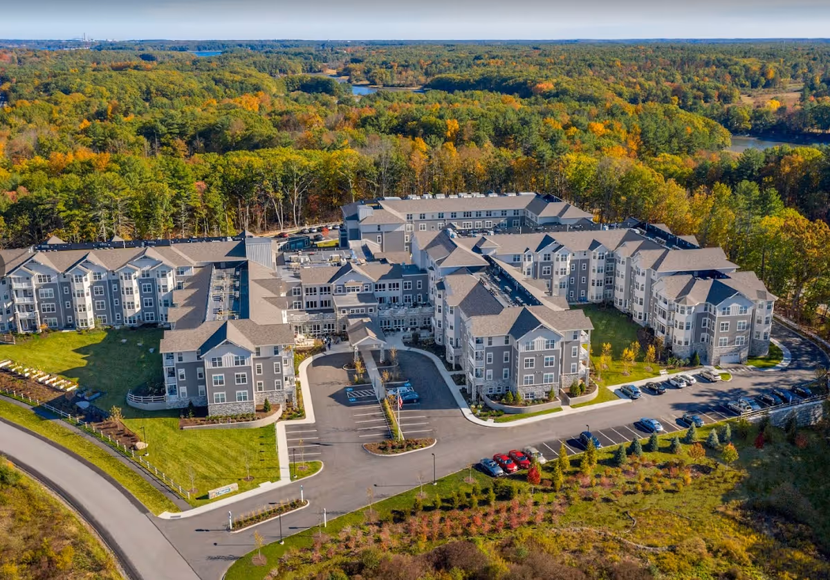 Aerial view of a large senior living facility named Riverwoods Durham surrounded by trees with autumn foliage. The multi-wing building has multiple stories with gray exterior walls and a large parking area in front. The facility is set in a green landscape with a road curving around the property.