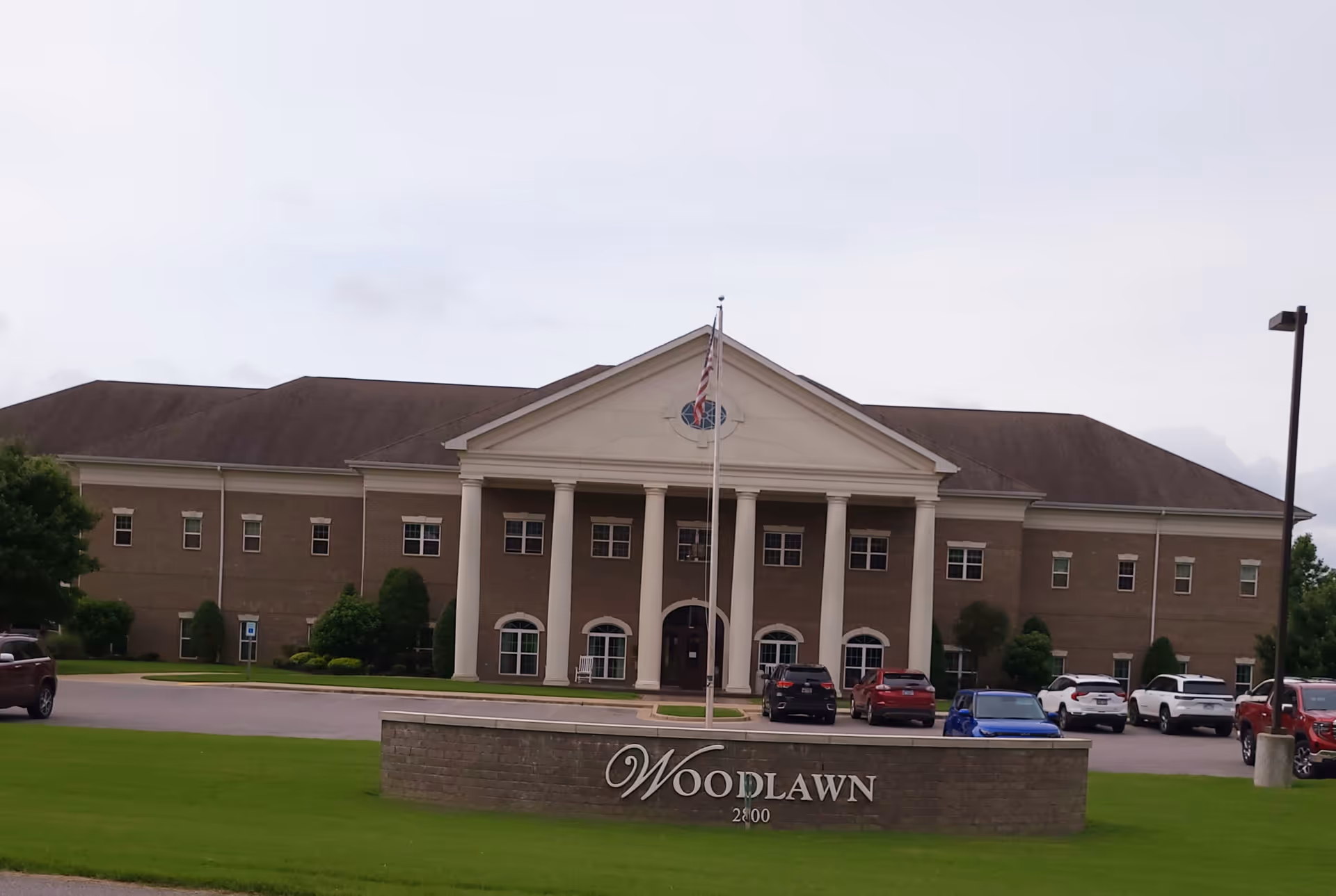 Front exterior view of a large two-story brick building with white columns and a triangular pediment. There is a flagpole with an American flag in front of the building, several parked cars, and a sign in the foreground that reads 'Woodlawn 2800'.