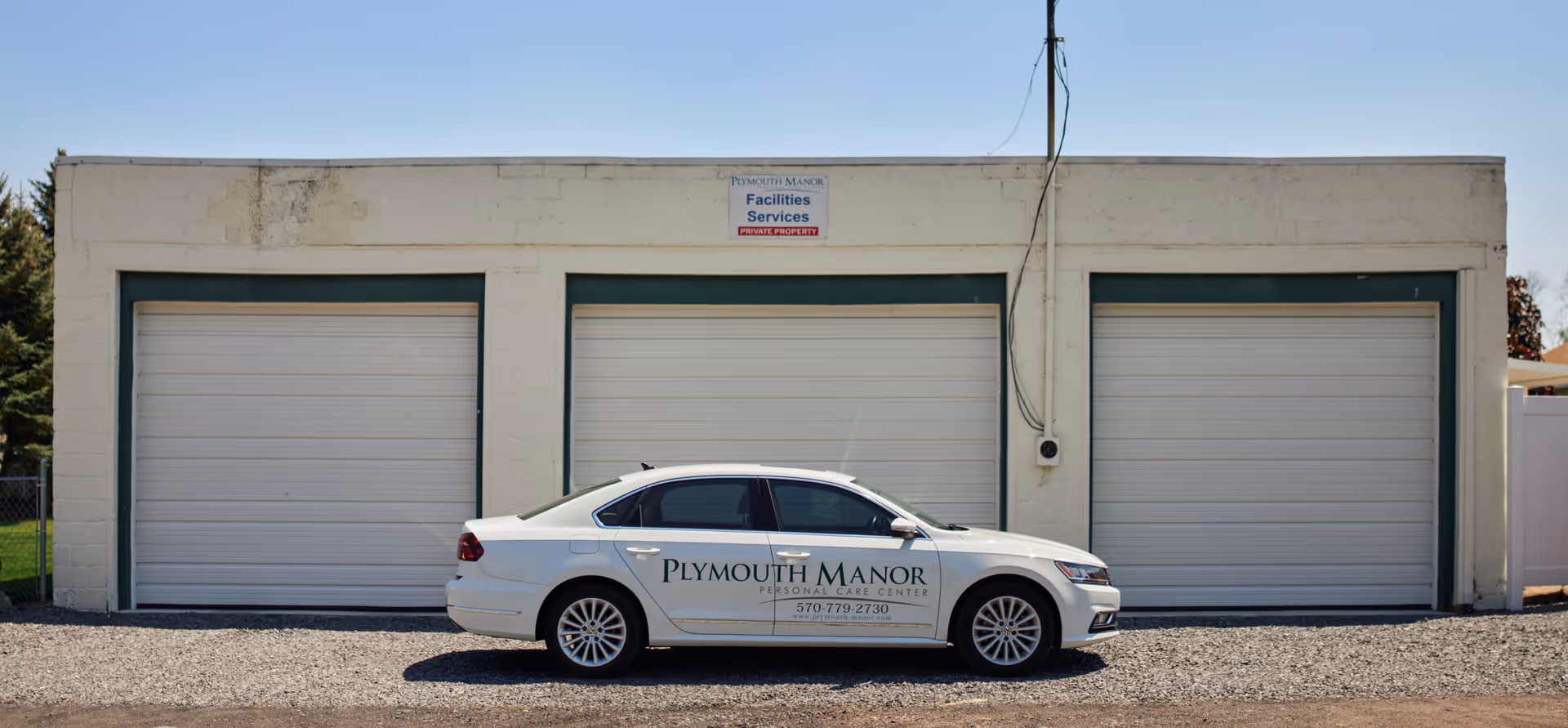 A white Plymouth Manor sedan parked in front of a three-bay garage building with closed roll-up doors.