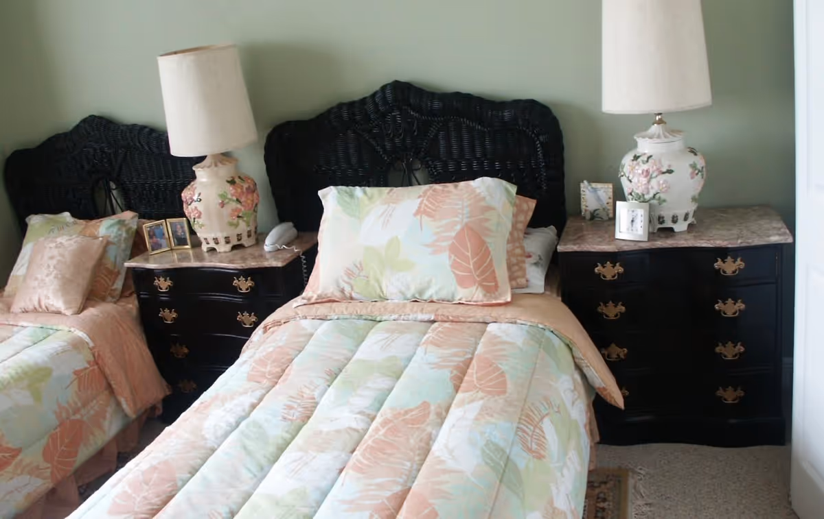 A bedroom with two twin beds featuring black wicker headboards and floral patterned bedding in pastel colors. Each bed has a nightstand with a decorative lamp, framed photos, and a clock. The walls are painted light green.