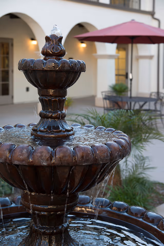 A close-up view of a decorative two-tiered water fountain with water flowing from the top tier to the bottom. In the background, there is a patio area with a table, chairs, a red umbrella, and a building with arched doorways and windows.