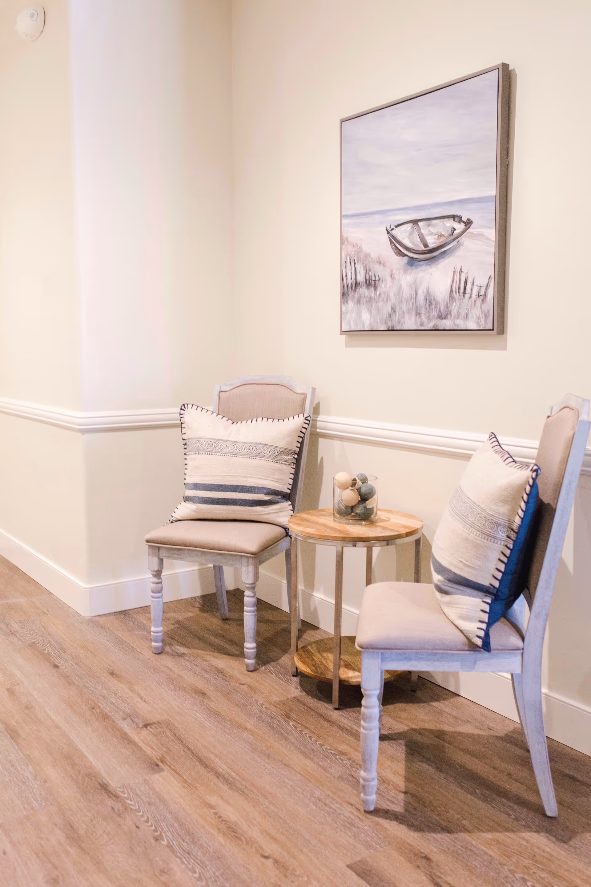 A cozy corner with two beige cushioned chairs featuring decorative pillows, a small round wooden side table with a glass container holding decorative balls, and a framed painting of a boat on a beach hanging on a light-colored wall.