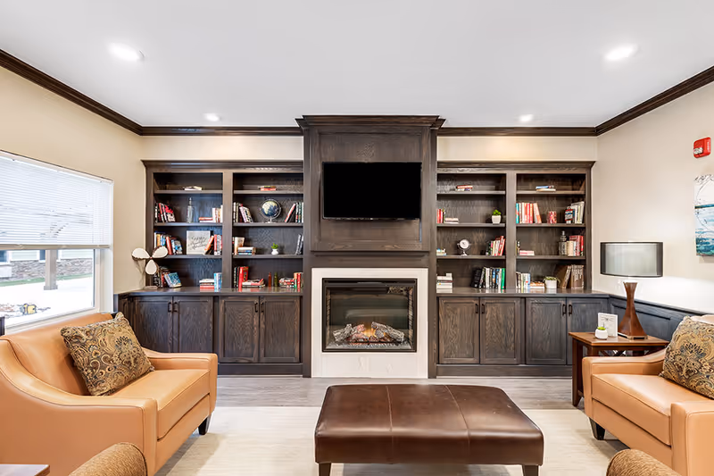 A cozy living room area with two tan leather armchairs facing a dark wood built-in bookshelf and fireplace. Above the fireplace is a mounted flat-screen TV. The shelves contain books, a globe, and decorative items. A large window with blinds is on the left, and a side table with a lamp is on the right. The room has light-colored walls and ceiling with dark wood trim.