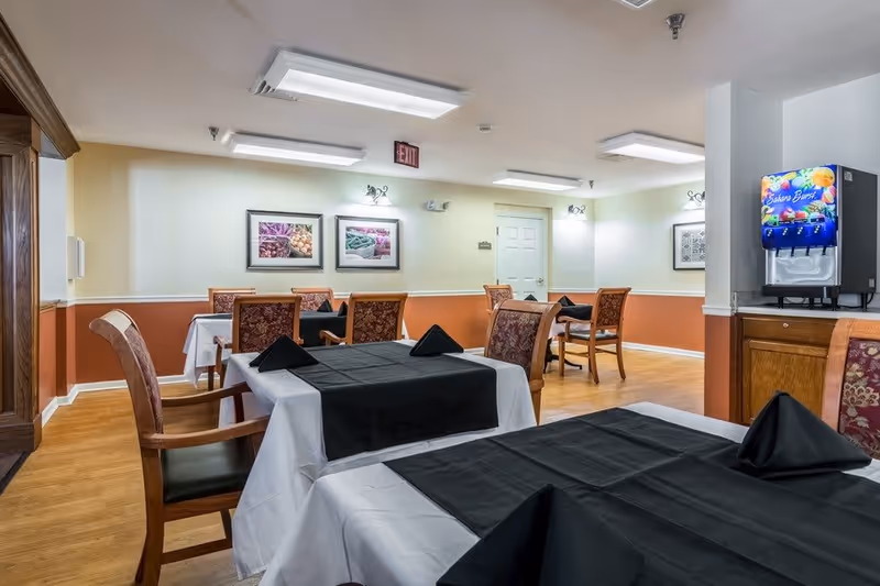 Interior view of a dining room in an assisted living facility with tables covered in white tablecloths and black runners, each set with black folded napkins. The room has wooden chairs with patterned upholstery, light-colored walls with a two-tone paint scheme, framed artwork on the walls, and a beverage dispenser on a wooden cabinet in the corner.