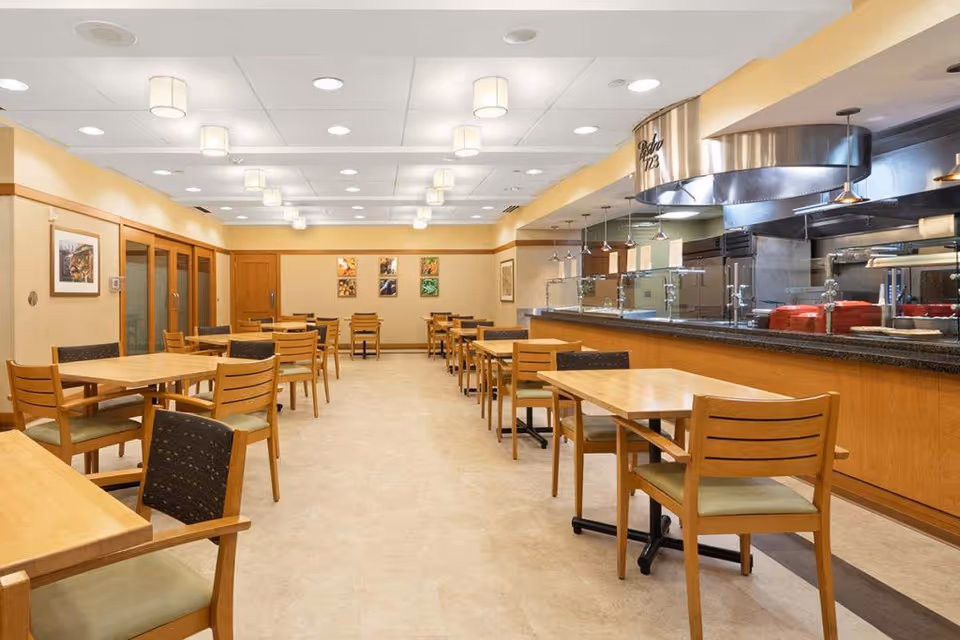 A clean and well-lit dining room with multiple wooden tables and chairs arranged neatly. The room features a serving counter with a stainless steel kitchen area behind it. The walls are decorated with framed pictures, and the ceiling has multiple round light fixtures.