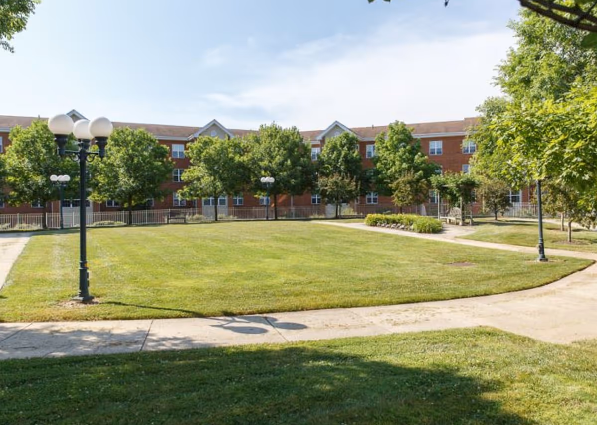 A well-kept grassy courtyard with walkways, lamp posts and trees in front of a three-story red brick building.