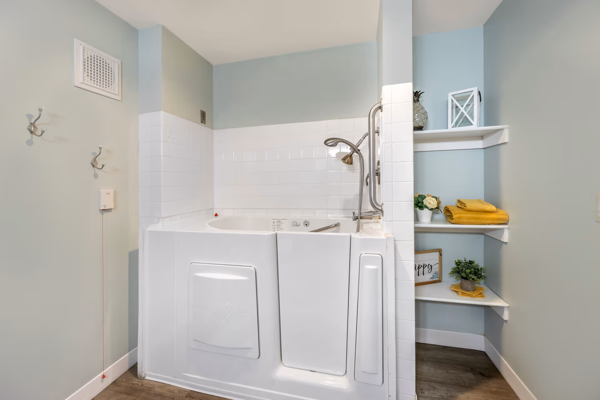 A walk-in bathtub with white tiled walls and a handheld showerhead in a bathroom. To the right of the tub are corner shelves holding folded yellow towels, a small potted plant, a decorative lantern, and a framed sign. The walls are painted light blue, and there are two metal hooks on the left wall.
