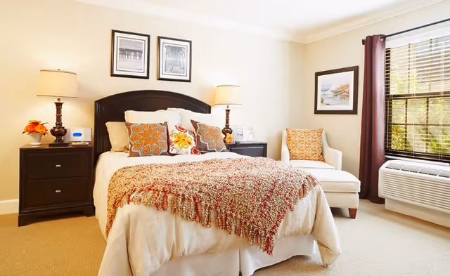 Bright, neatly made bedroom with a central bed topped with patterned pillows and a fringed throw, flanked by nightstands with lamps, an armchair by a window, and a wall air-conditioning unit.