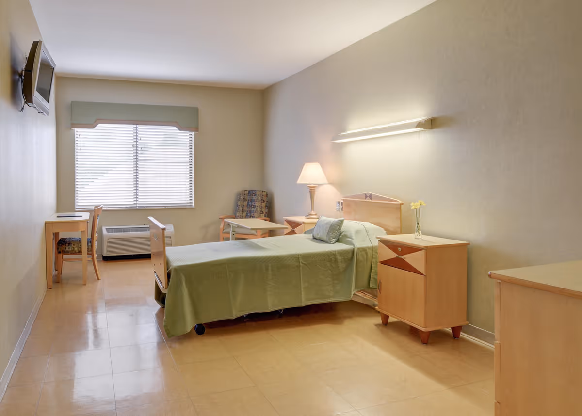 A simple, clean bedroom in a senior living facility with a single bed covered in a green bedspread, a bedside table with a lamp and a small vase with flowers, a chair with a small table, a desk with a chair, a wall-mounted TV, and a window with blinds letting in natural light.
