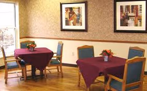 A dining area with purple tablecloths, framed wall art, and wooden chairs in a care facility.