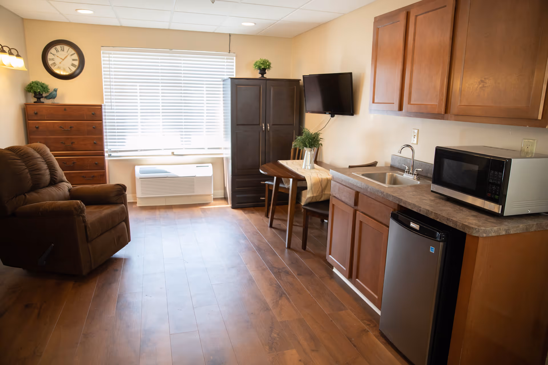 A cozy assisted living room with wooden flooring, a brown recliner chair, a wooden dresser with a plant and bird decoration, a wall clock, a window with blinds, a dark wooden cabinet, a small dining table with two chairs and a plant centerpiece, a wall-mounted TV, and a kitchenette area with wooden cabinets, a sink, a microwave, and a mini refrigerator.