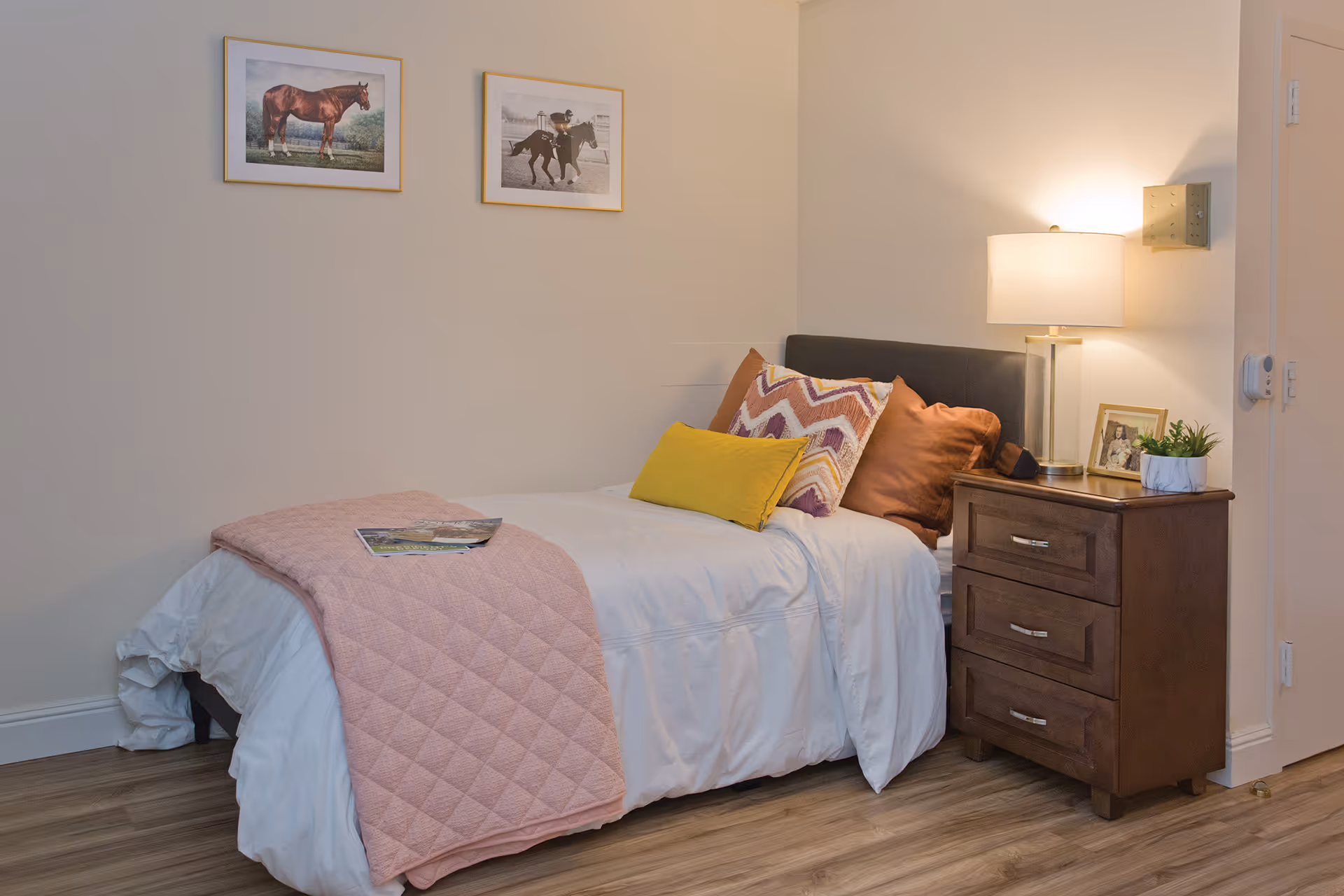 A neatly made single bed with white bedding, a pink quilted blanket, and three decorative pillows in yellow, brown, and patterned colors. Next to the bed is a wooden nightstand with three drawers, a table lamp, a framed photo, and a small potted plant. Two framed pictures of horses hang on the beige wall above the bed. The room has wooden flooring.