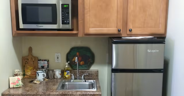 Small kitchenette with a microwave and wooden cabinets above a sink and countertop holding mugs and decor, and a stainless steel mini fridge.
