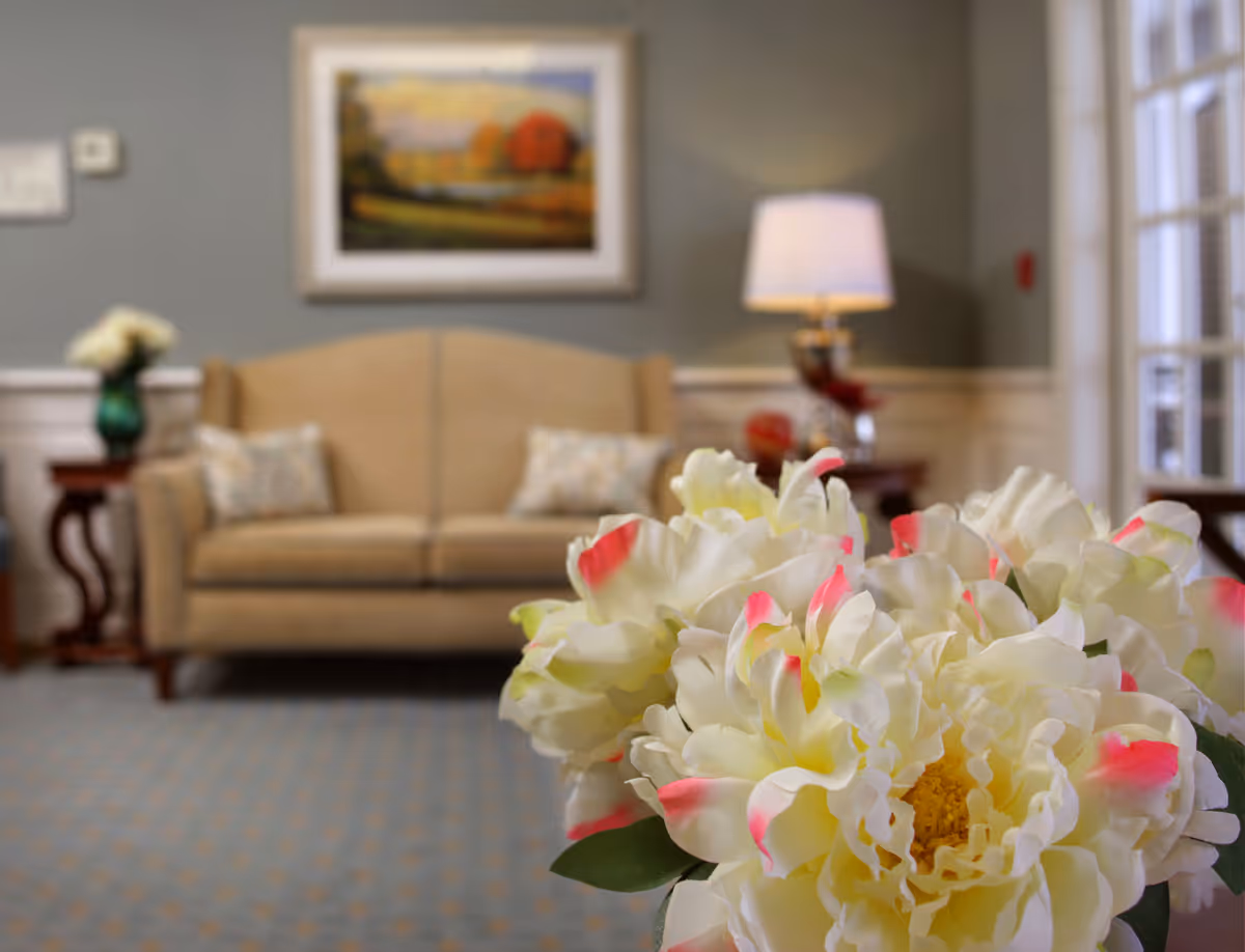 Close-up of a bouquet of white flowers with pink tips in the foreground, with a beige sofa, side tables, a lamp, and a framed landscape painting in a softly lit living room in the background.