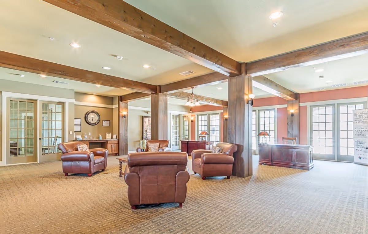 A spacious and well-lit common area with beige carpet and wooden beams on the ceiling. The room features several brown leather armchairs arranged around a wooden coffee table. There are large windows and glass doors letting in natural light, and a wooden reception desk is visible in the background. The walls are painted in warm tones with some decorative elements including a clock and framed items.