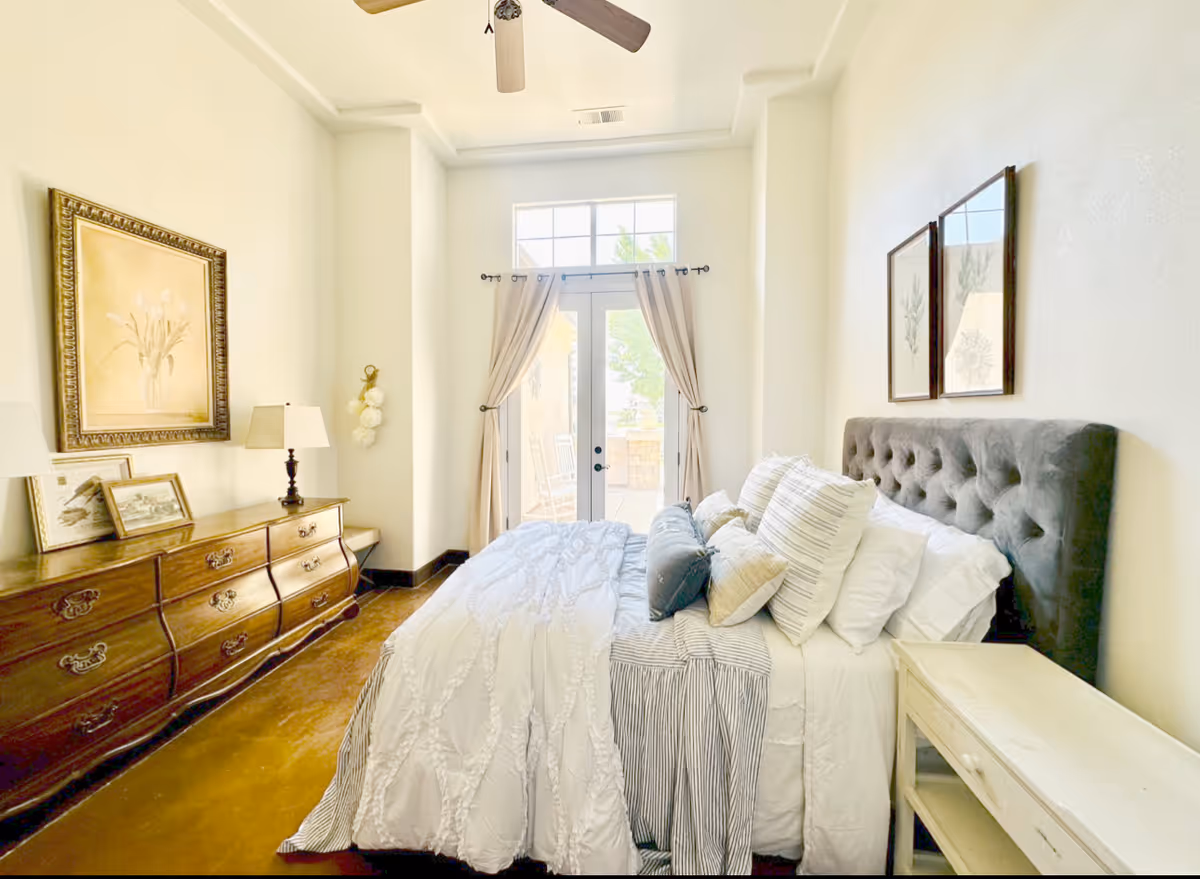 Sunlit bedroom with a tufted headboard and neatly made bed, wooden dresser, and French doors with curtains leading outside.