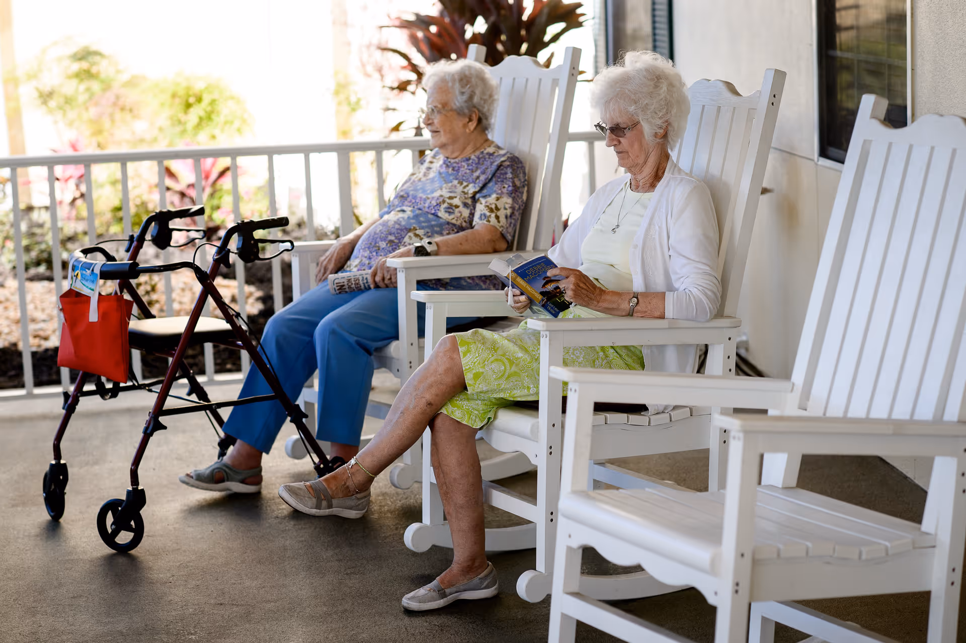 Two elderly women sitting on white wooden rocking chairs on a porch. One woman is reading a book while the other is holding a newspaper. A walker with a red bag is positioned next to one of the women. There are plants and a railing in the background.