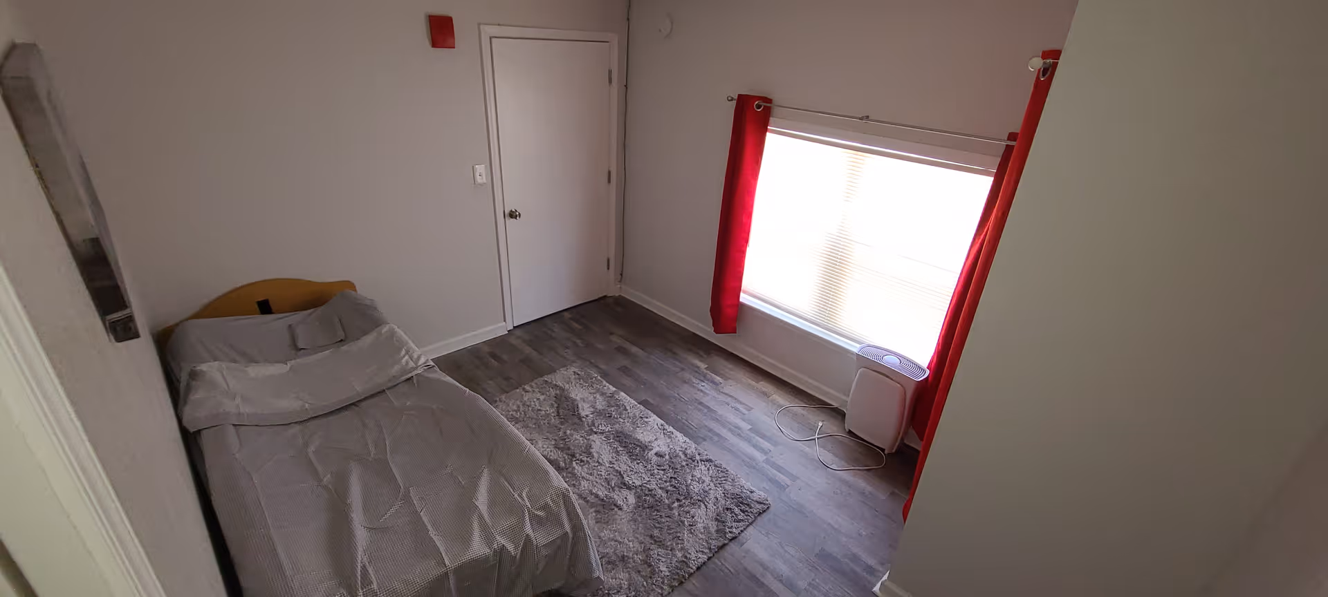 Small bedroom with a single bed, gray bedding, a shag rug on wood-look floors, a window with red curtains and a small air purifier next to a closed door.