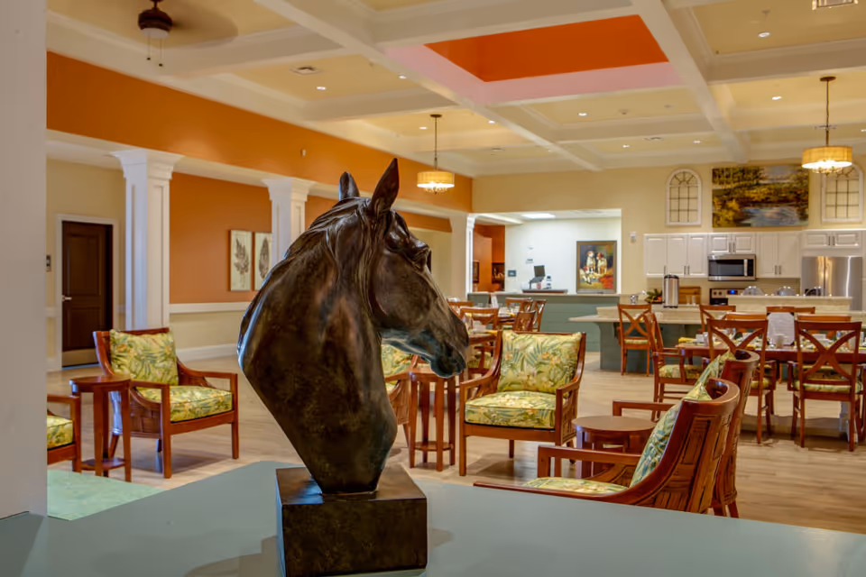 Interior view of a senior living facility common area featuring a bronze horse head sculpture in the foreground. The room has wooden chairs with green floral cushions, tables, and a kitchen area with white cabinets, a microwave, and a refrigerator in the background. The ceiling has recessed lighting and ceiling fans, with warm orange and beige walls.