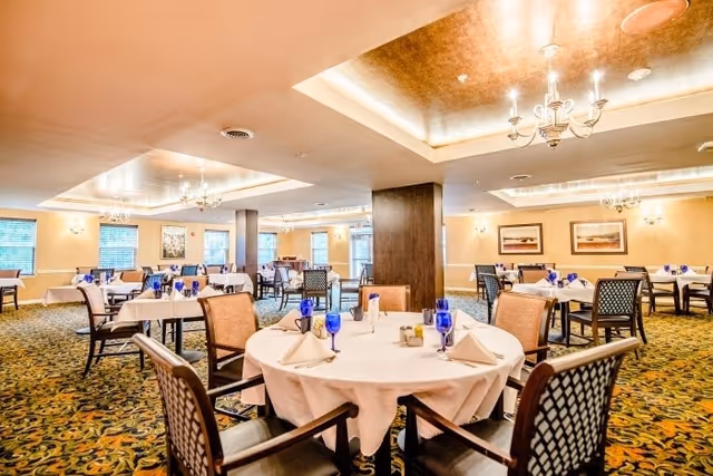 A spacious dining room with multiple round and rectangular tables covered with white tablecloths. Each table is set with blue water glasses, folded white napkins, and silverware. The room has patterned carpet flooring, beige walls, and large windows letting in natural light. Elegant chandeliers hang from the ceiling, and framed artwork decorates the walls.