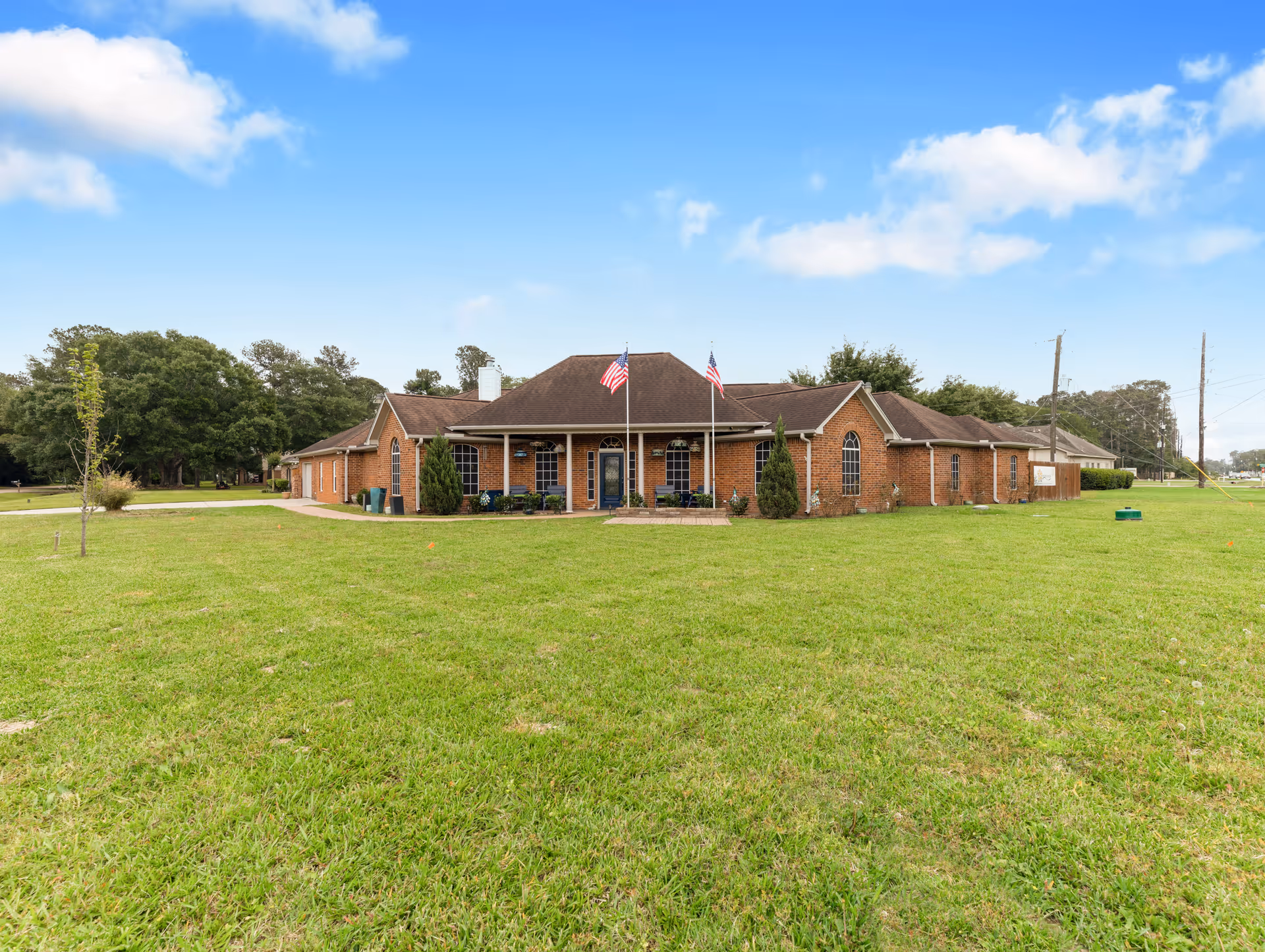 Front exterior view of a single-story brick building with a large green lawn in front. The building has a dark roof, several windows, two American flags on flagpoles near the entrance, and some small bushes and plants around the entrance area. The sky is partly cloudy.
