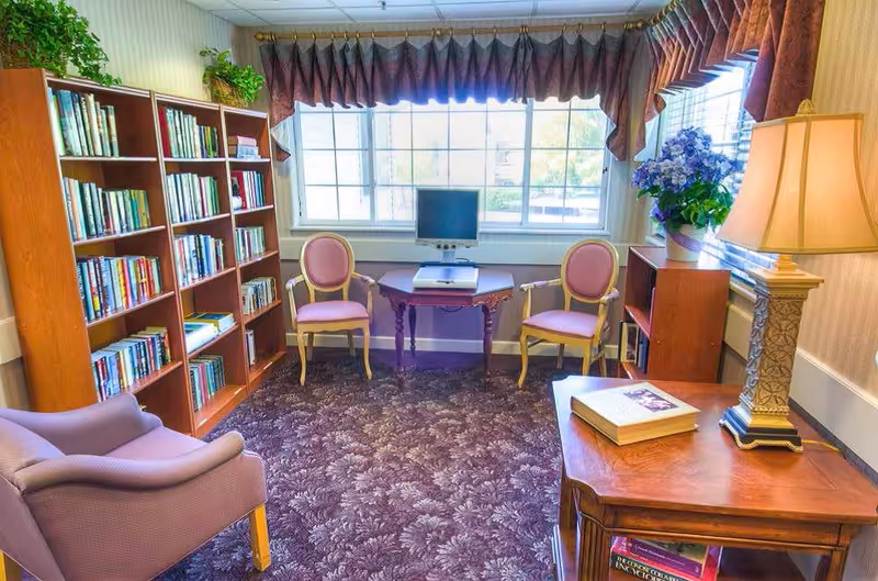 Cozy reading room with bookshelves, upholstered chairs, a small table with a computer, and a lamp by a large window.