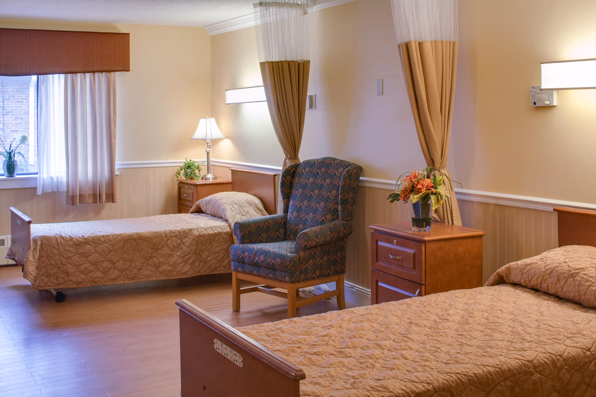 A well-lit room with two single beds covered in beige quilts, a patterned armchair between them, wooden nightstands with a lamp and a flower vase, beige walls, and a window with sheer curtains and a brown valance.