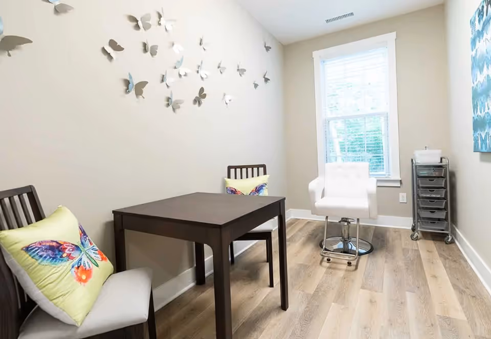 A small, bright room with light-colored walls and wood flooring. There is a dark wooden table with two wooden chairs, each with a colorful butterfly pillow. On the wall above the table, there are decorative 3D butterfly wall hangings. A white cushioned swivel chair is positioned near a window with white blinds. A small metal cart with drawers is placed against the wall near the window.