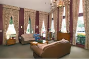 A cozy living room with tall windows covered by patterned curtains, two sofas facing each other with a small table in between, a wooden cabinet, and a person sitting on one of the sofas. The room has a chandelier and framed pictures on the walls.