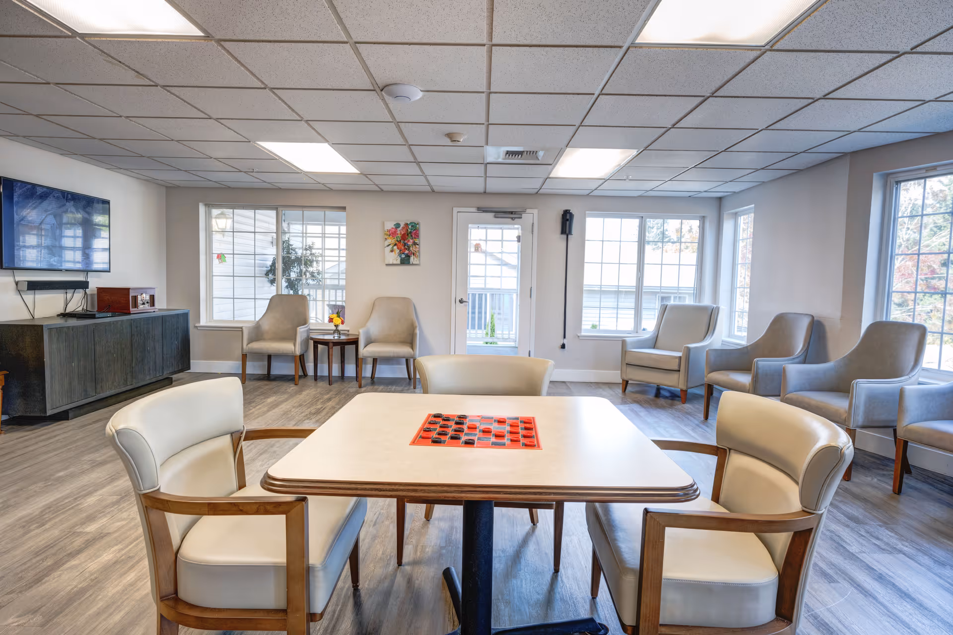 A bright and spacious common area with a square table in the center featuring a checkers game board. Four cushioned chairs with wooden armrests surround the table. The room has large windows letting in natural light, several additional cushioned chairs along the walls, a flat-screen TV mounted on the wall above a dark wooden cabinet, and a small table with a flower vase between two chairs near the windows.