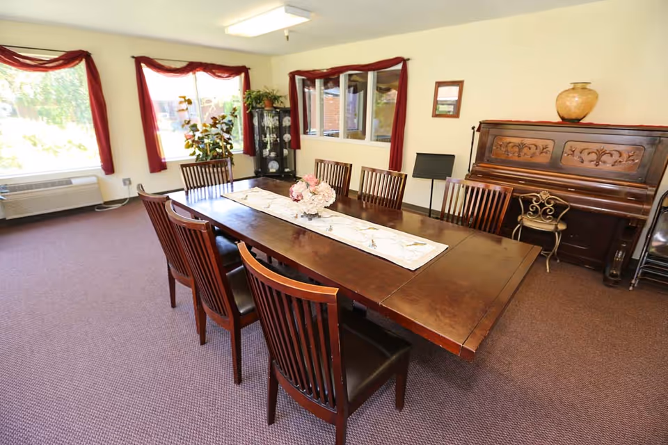 A long wooden dining table surrounded by chairs in a bright common room with windows, red curtains, plants, and an upright piano.