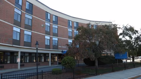 Exterior view of a multi-story brick building with several windows and a curved facade. There is a tree and some bushes in front of the building, along with a black metal fence and a sidewalk. A blue sign is visible near the entrance, and a streetlamp stands near the sidewalk.
