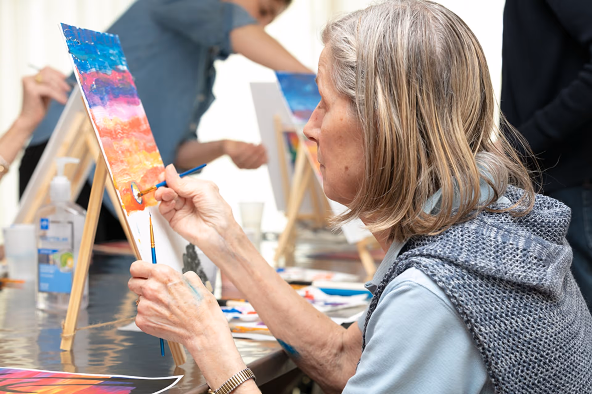 An elderly woman painting a colorful sunset scene on a canvas at an easel during an art activity, with other participants painting in the background.