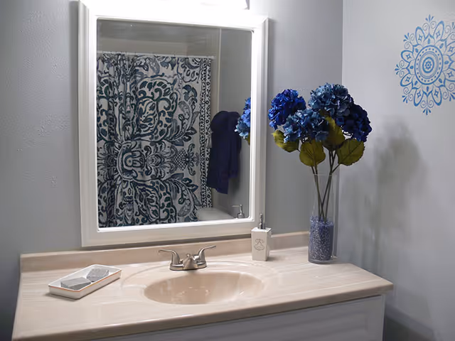 A bathroom vanity with a beige countertop, an oval sink, and a silver faucet. Above the sink is a white-framed mirror reflecting a shower curtain with a blue and white intricate pattern. To the right of the sink is a clear vase with blue flowers and green leaves, and a soap dispenser. The wall is light gray with a blue decorative decal.