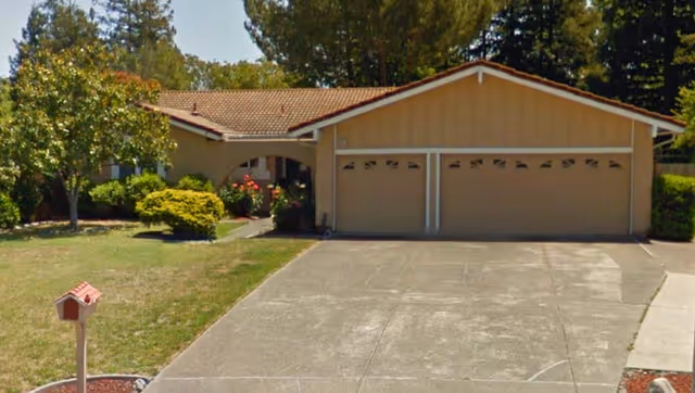 Single-story residential building with a two-car garage, a driveway, and a front yard featuring a mailbox, bushes, trees, and flowers.