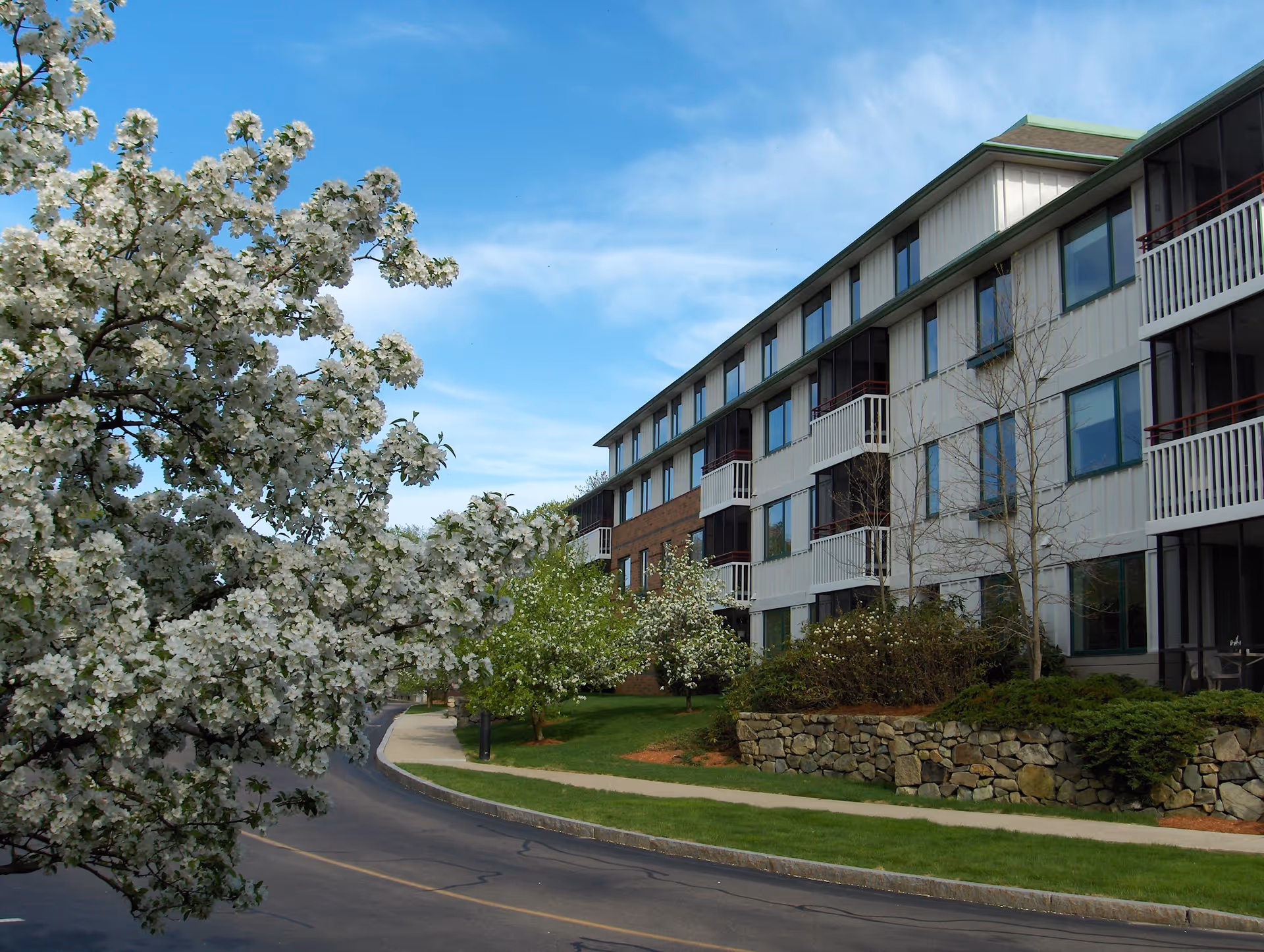 Exterior view of a multi-story senior living facility building with balconies, surrounded by landscaped greenery including flowering trees and bushes, under a blue sky with some clouds.