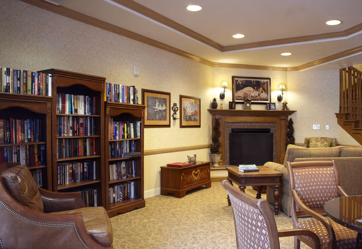 A cozy living room area with wooden bookshelves filled with books, a leather armchair, a wooden coffee table, patterned upholstered chairs around a glass-top table, a fireplace with a framed picture above it, and warm lighting from wall sconces and recessed ceiling lights.