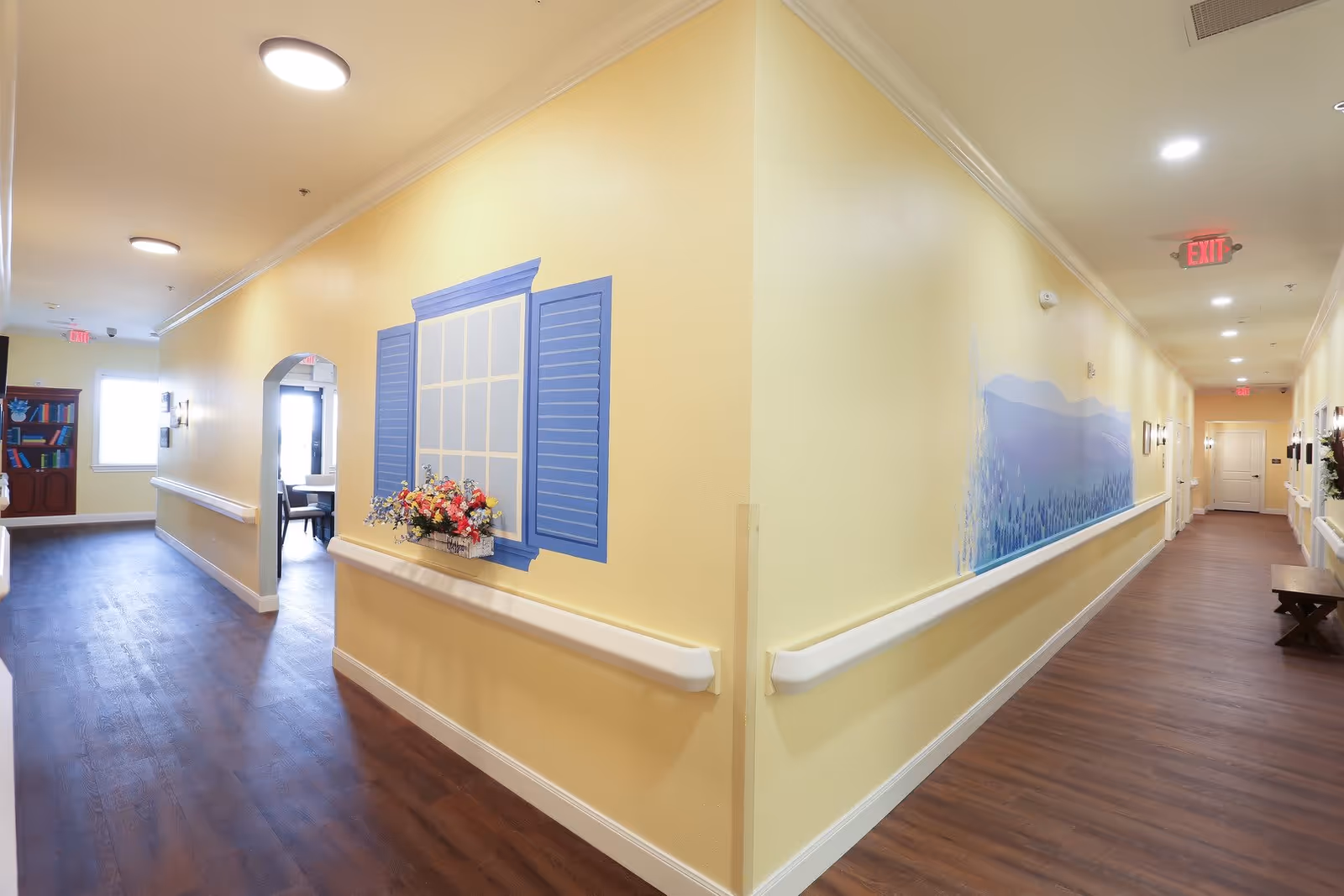A bright, clean hallway in Serenity Oaks Assisted Living and Memory Care with yellow walls, wood flooring, and white handrails on both sides. The left wall features a painted window with blue shutters and a flower box with colorful flowers. The right wall has a mural of blue mountains. The hallway is well-lit with ceiling lights and has exit signs visible at both ends. There is a small wooden bench on the right side near the end of the hallway.