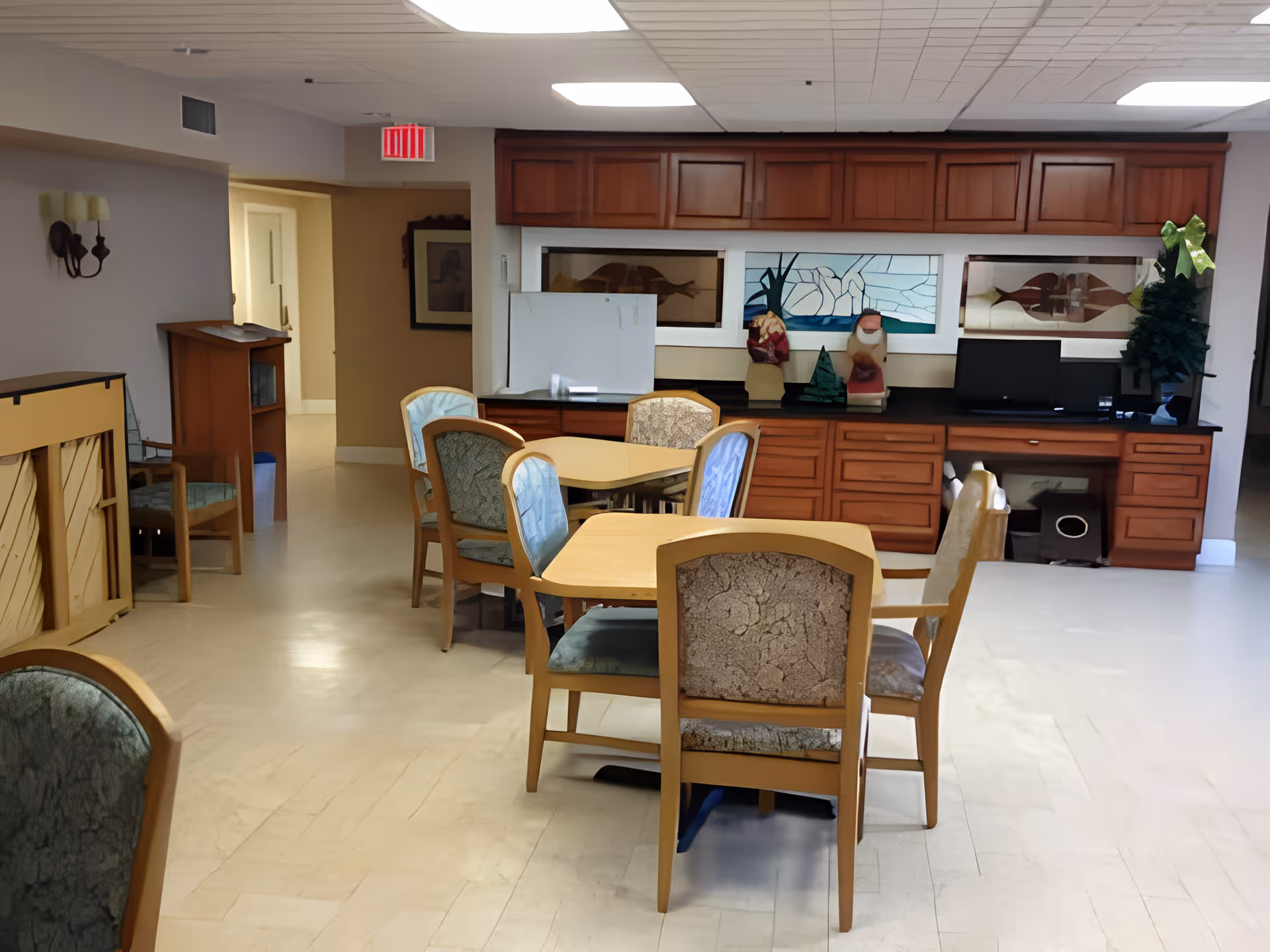 Interior room with several wooden tables and chairs arranged in the center. The back wall features wooden cabinets with decorative stained glass panels and various ornaments. The floor is light-colored, and there is an exit sign above a doorway on the left side.