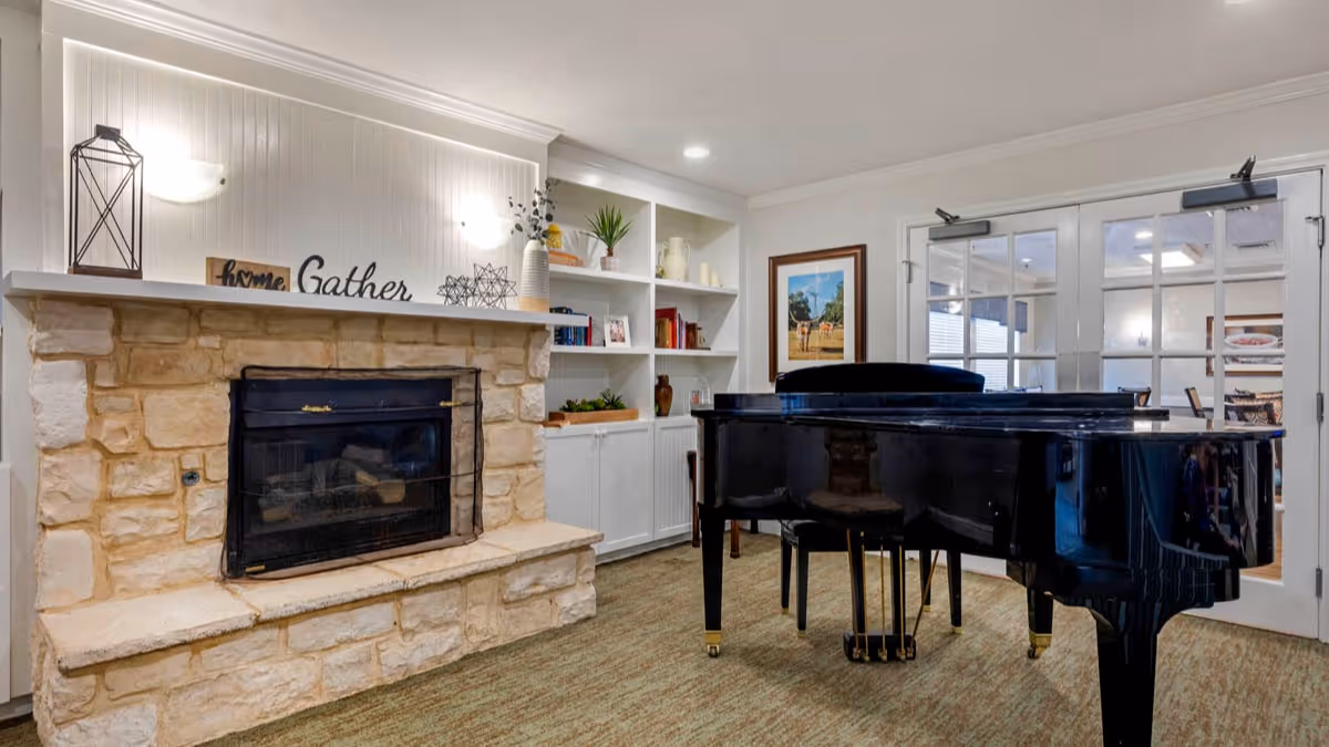 A lounge featuring a stone fireplace with a mantel and built-in shelves, and a black grand piano near glass double doors.