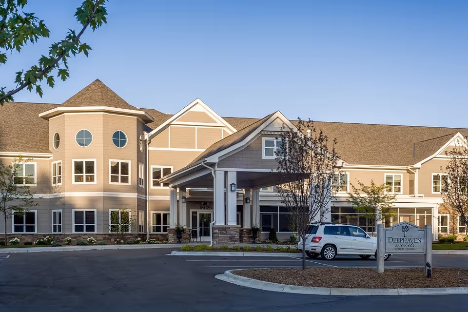 Exterior view of Deephaven Woods Senior Living facility showing a large, beige building with multiple windows, a covered entrance, a parked white SUV, and a sign with the facility name. The sky is clear and there are some trees and landscaping around the building.