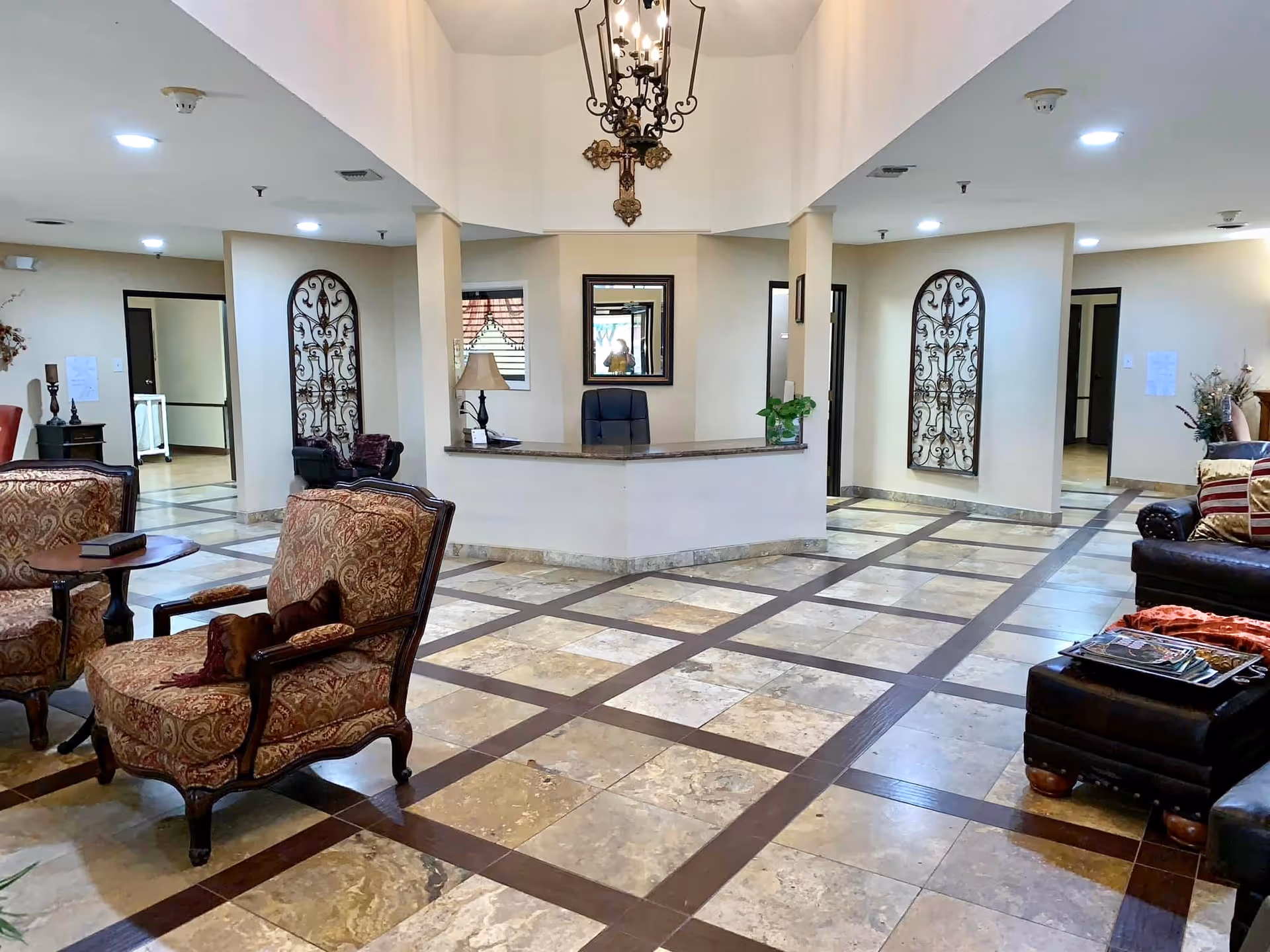 Interior view of a senior living facility lobby area with a reception desk in the center, two patterned armchairs and a small round table on the left, and a dark leather sofa with a matching ottoman on the right. The floor has a tiled pattern with brown and beige colors. Decorative wrought iron wall hangings and a chandelier are visible, along with several doorways leading to other rooms.