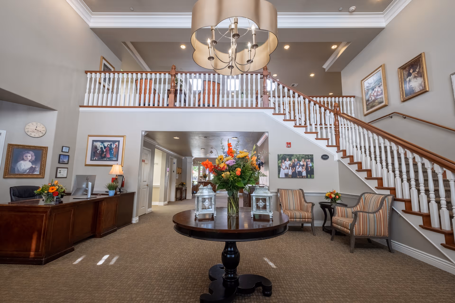 A spacious and well-lit interior lobby area of a senior living facility featuring a round wooden table with a large floral arrangement and two decorative lanterns. To the left is a wooden reception desk with a computer and flowers. On the right, there are two striped armchairs with a small side table holding a flower arrangement. A staircase with white spindles and wooden handrails leads to an upper level with additional seating. The walls are adorned with framed artwork and photographs, and a large modern chandelier hangs from the ceiling.
