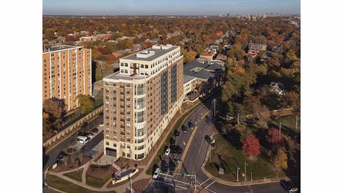 Aerial view of a multi-story senior living facility building surrounded by roads, trees with autumn foliage, and nearby residential buildings under a clear sky.