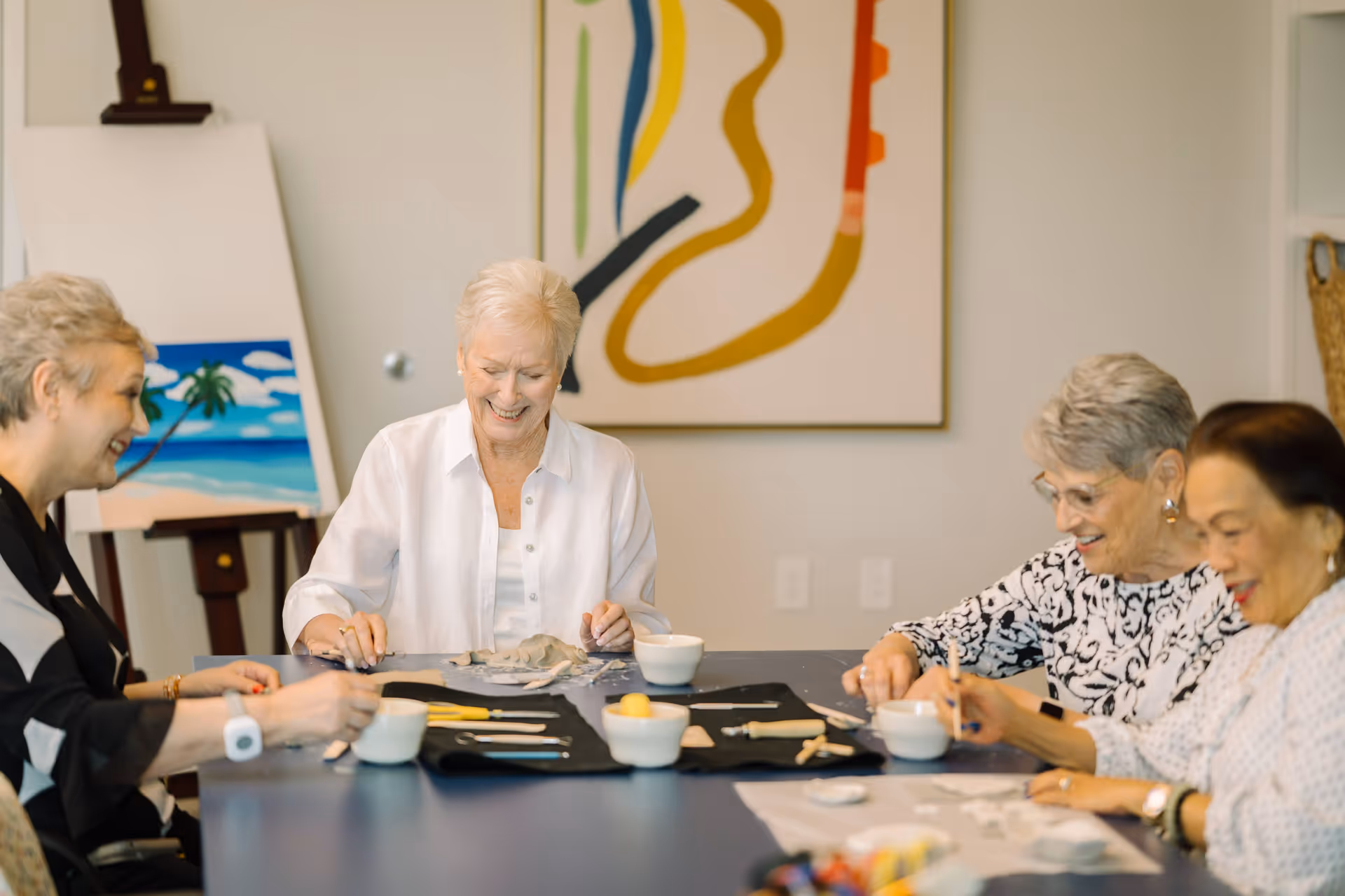 Four elderly women sitting around a table engaged in an arts and crafts activity, smiling and enjoying each other's company. There are art supplies and small bowls on the table. In the background, there is a colorful abstract painting on the wall and an easel with a painting of a beach scene.