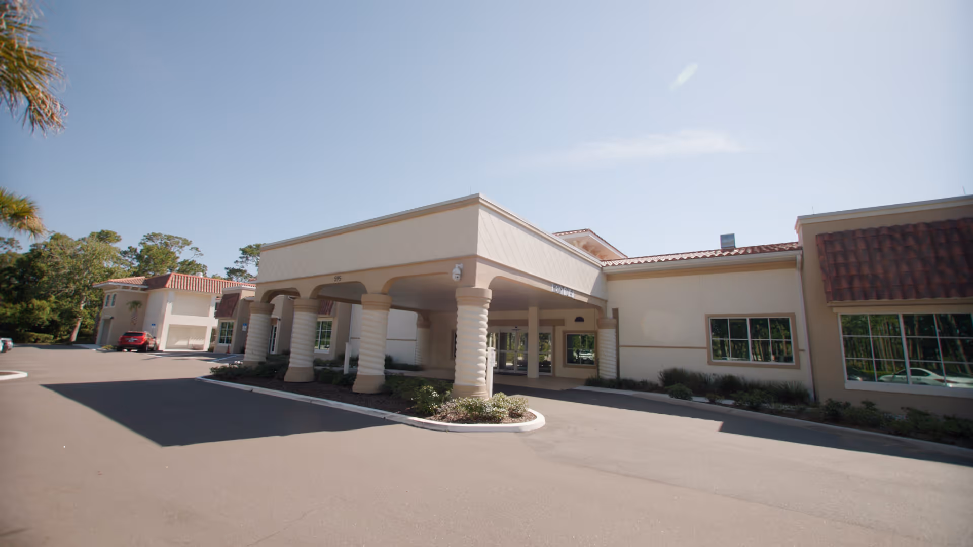 Exterior view of a single-story nursing and rehabilitation facility with a covered entrance supported by columns, surrounded by a paved driveway and landscaped greenery under a clear sky.
